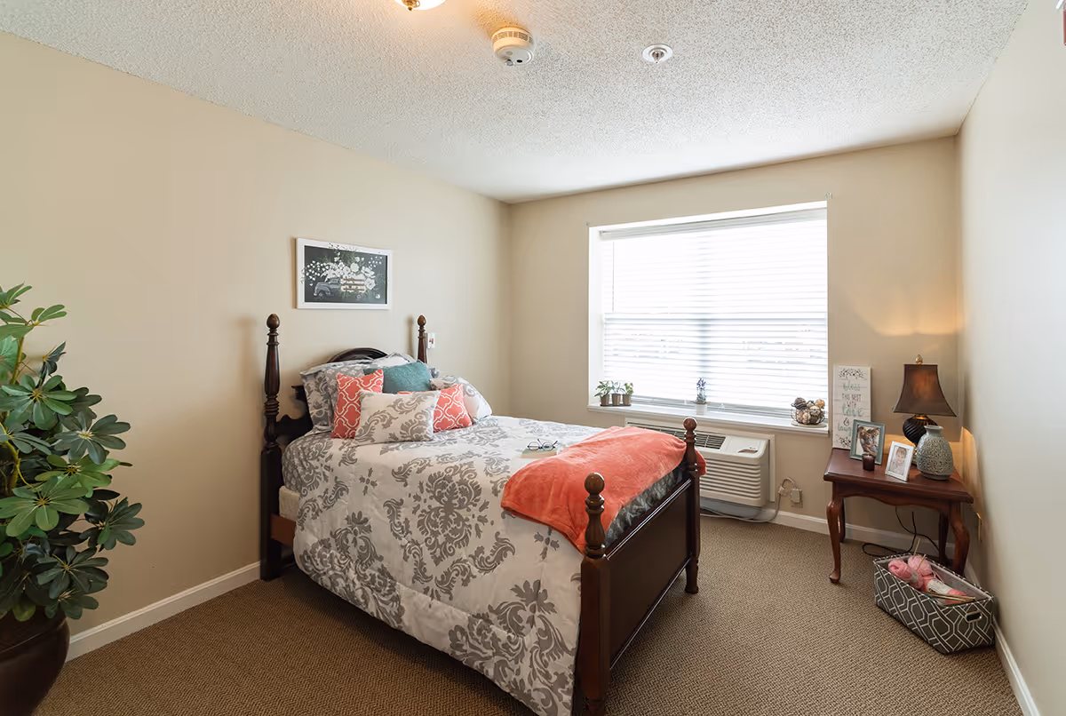 Well-lit furnished bedroom with a wooden bed dressed in patterned bedding, a bedside table with lamp and photos, a window air conditioner, and a potted plant.
