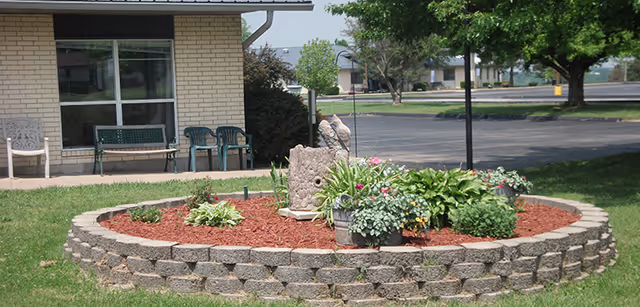 A circular raised garden bed made of stone blocks filled with mulch, various green plants, and flowers. Behind the garden bed is a building with a window and several chairs placed outside on a concrete patio. Trees and a parking lot are visible in the background.