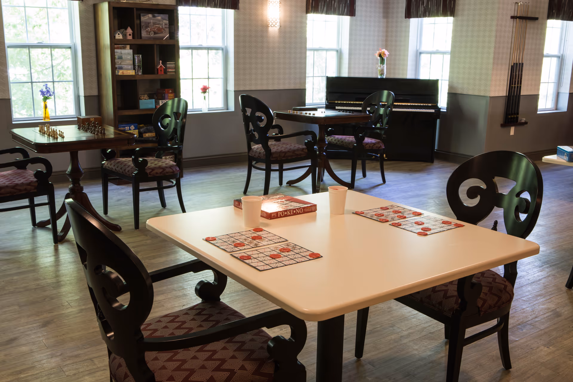 A well-lit common room with several tables and chairs arranged for games and social activities. One table in the foreground has bingo cards and two cups, while another table has a chessboard. A bookshelf with board games and decorative items is visible near large windows. A piano stands against the far wall with a vase of flowers on top. The room has wooden flooring and patterned wallpaper.