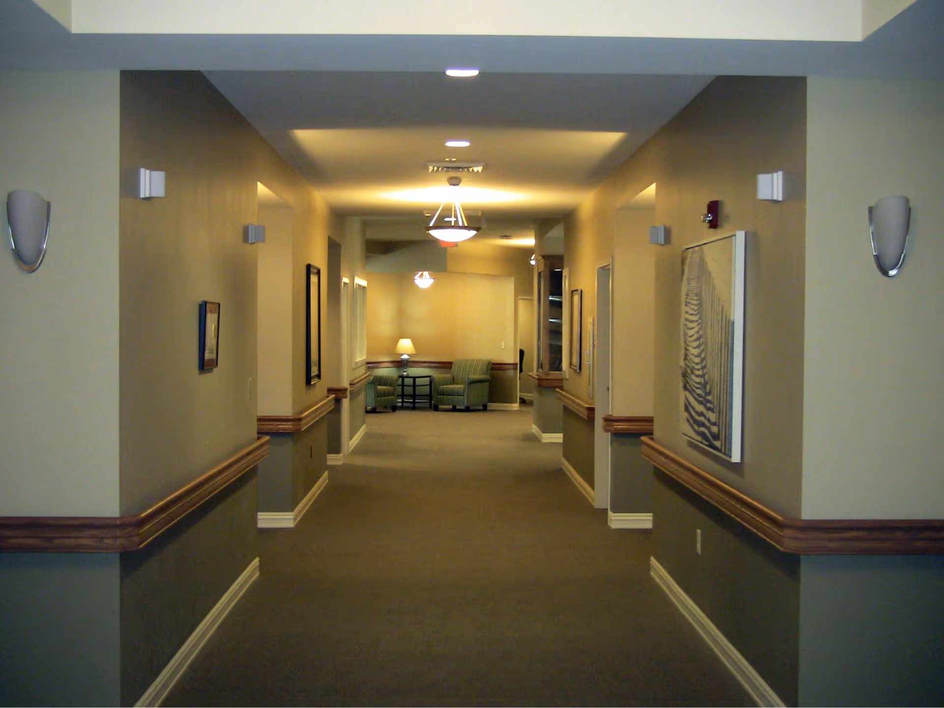 A long, carpeted hallway in a senior living facility with beige and light green walls, wooden chair rails, wall-mounted lights, framed artwork, and a seating area with two green armchairs and a small table with a lamp at the far end.