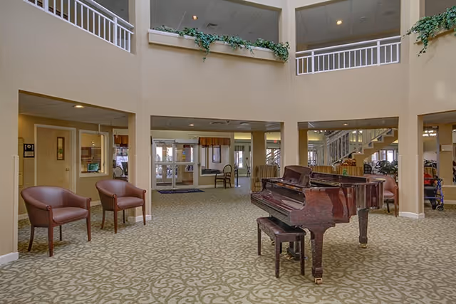 Spacious senior living facility common area with a grand piano in the center, two brown chairs on the left, beige patterned carpet, and a two-story open ceiling with plants on the upper ledge.