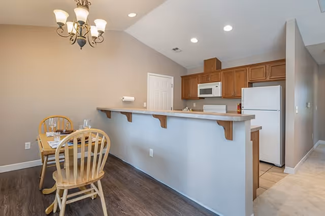 Interior view of a kitchen and dining area in a senior living facility. The dining area has a wooden table set with plates, glasses, and cutlery, accompanied by wooden chairs. The kitchen features wooden cabinets, a white refrigerator, a white microwave, and a countertop with a raised bar supported by decorative brackets. The walls are painted beige, and the floor is a combination of dark wood and tile. A chandelier with multiple lights hangs above the dining table.