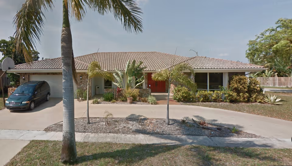 Front exterior view of a single-story house with a tiled roof, a driveway with a parked green minivan, palm trees, and various shrubs and plants in the front yard under a clear sky.