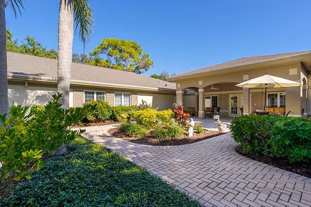 Outdoor courtyard area of a senior living facility with a paved walkway, green shrubs, palm trees, and a covered patio with seating and an umbrella under a clear blue sky.