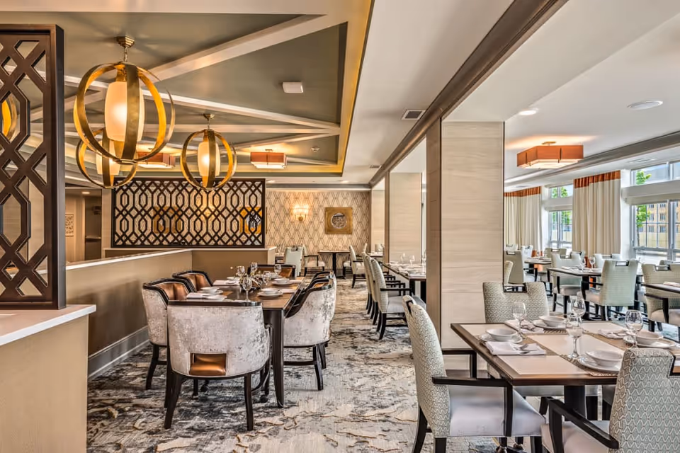 A spacious and elegantly decorated dining room with multiple tables set with plates, glasses, and napkins. The room features modern light fixtures with gold accents, patterned carpet, and large windows with white curtains allowing natural light to fill the space.