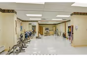 A spacious rehabilitation therapy room with exercise equipment including stationary bikes, parallel bars for walking practice, and various physical therapy tools along the walls. The room has a white tiled floor, beige walls with a decorative border near the ceiling, and fluorescent lighting.