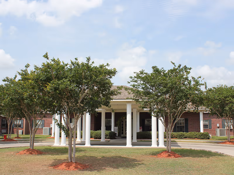 Front exterior view of a single-story brick building with white columns at the entrance, surrounded by green trees and a circular driveway under a partly cloudy sky.