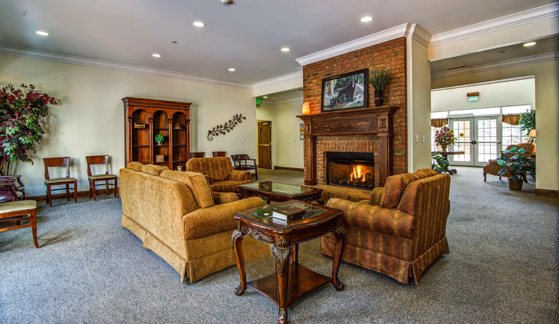 A cozy living room area in a senior living facility featuring a brick fireplace with a fire burning, surrounded by comfortable upholstered sofas and armchairs. There is a wooden coffee table and side table with decorative items. The room has carpeted floors, a wooden bookshelf, several chairs along the wall, and plants adding greenery. Large windows and glass doors in the background allow natural light to enter the space.