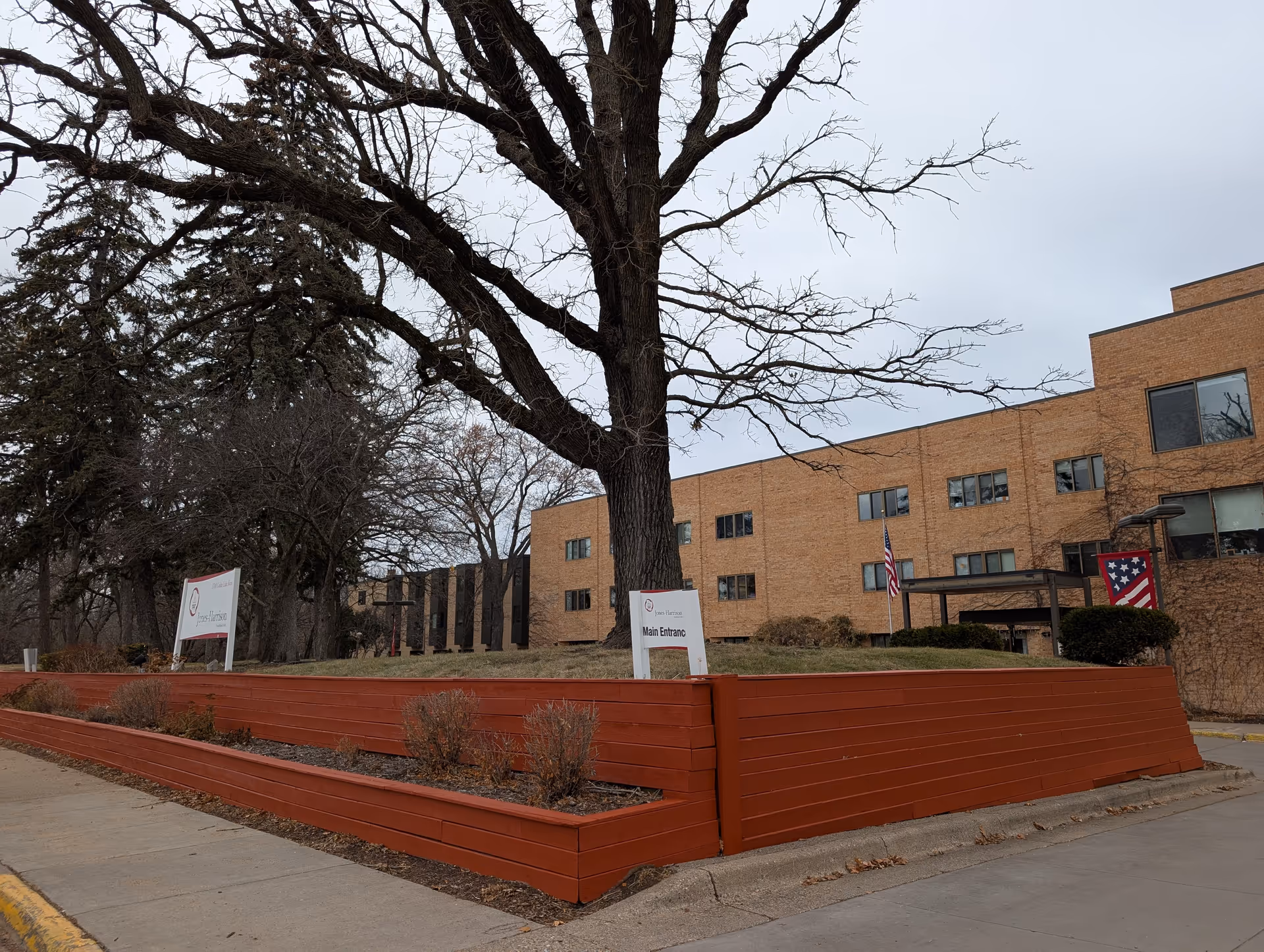 Front exterior of a brick senior living building with a large leafless tree, red raised planters, and a 'Main Entrance' sign.