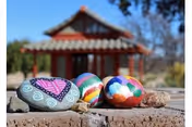 Close-up of four colorful painted rocks placed on a brick surface with a traditional-style building with a tiled roof and wooden beams blurred in the background under a clear blue sky.