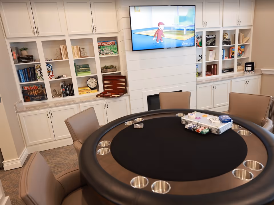 Round poker table with cup holders and chips surrounded by chairs in a communal room with built-in white shelving and a wall-mounted TV.