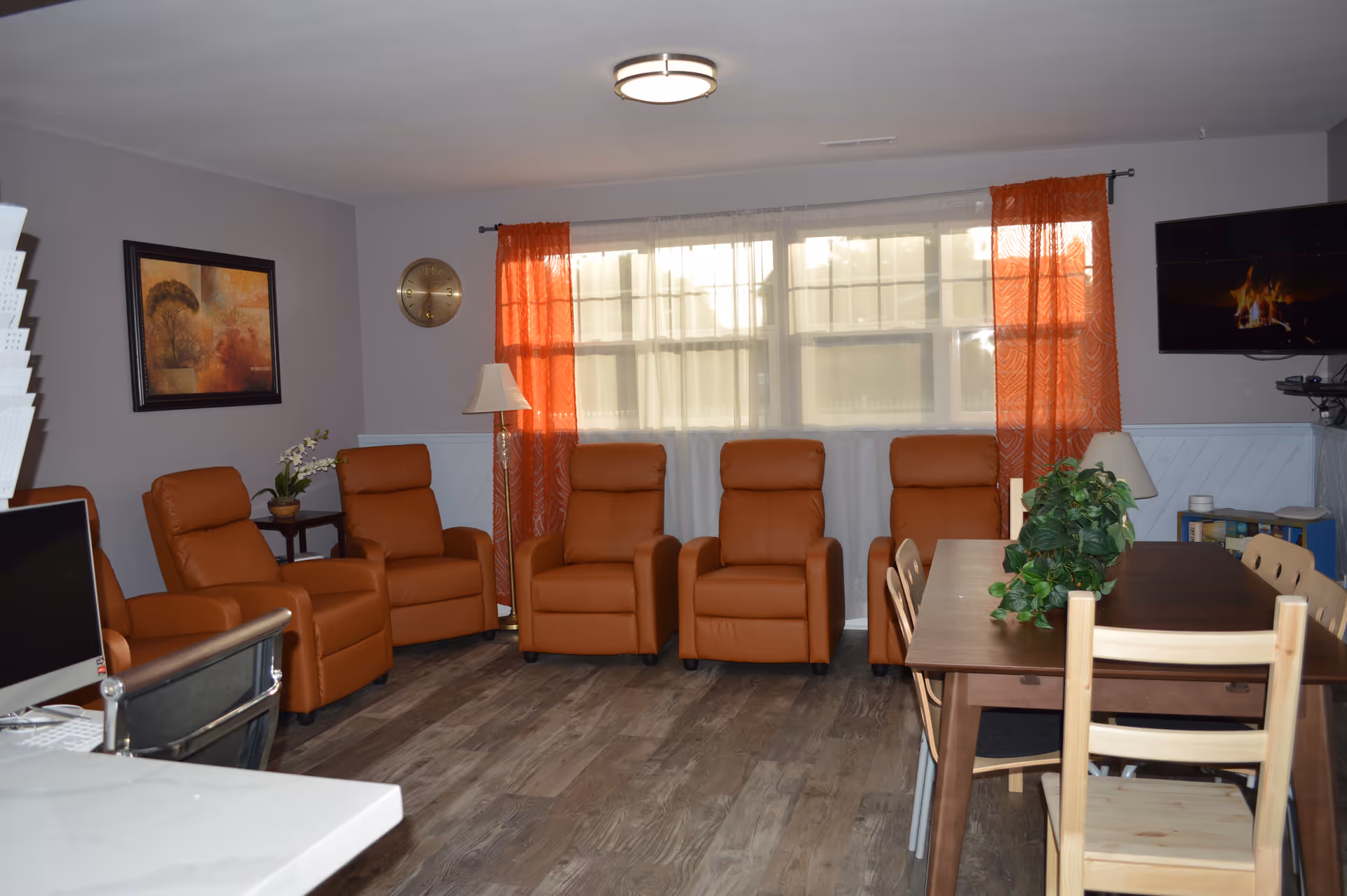 A cozy living room with six brown leather recliners arranged in a row against a wall with a large window covered by sheer white and orange curtains. There is a wooden dining table with chairs and a green plant centerpiece. A flat-screen TV mounted on the wall displays a fireplace scene. The room has wood flooring, a wall clock, a floor lamp, and a framed painting.