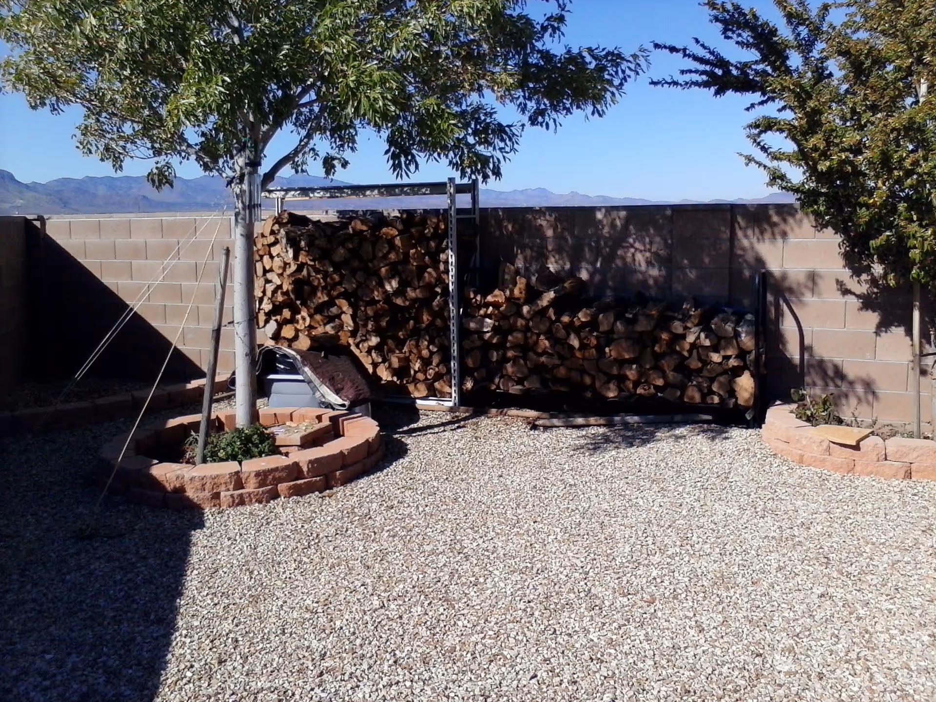 Outdoor area with gravel ground, two small trees planted in circular brick planters, and a large stack of firewood against a brick wall under a clear blue sky with mountains in the background.