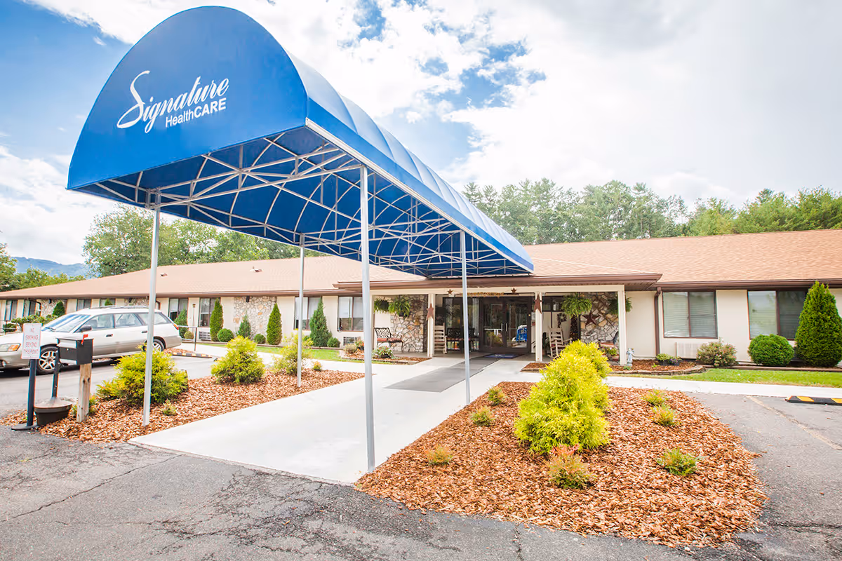 Entrance of a single-story care facility with a large blue canopy reading "Signature HealthCARE" over the driveway and landscaped beds.