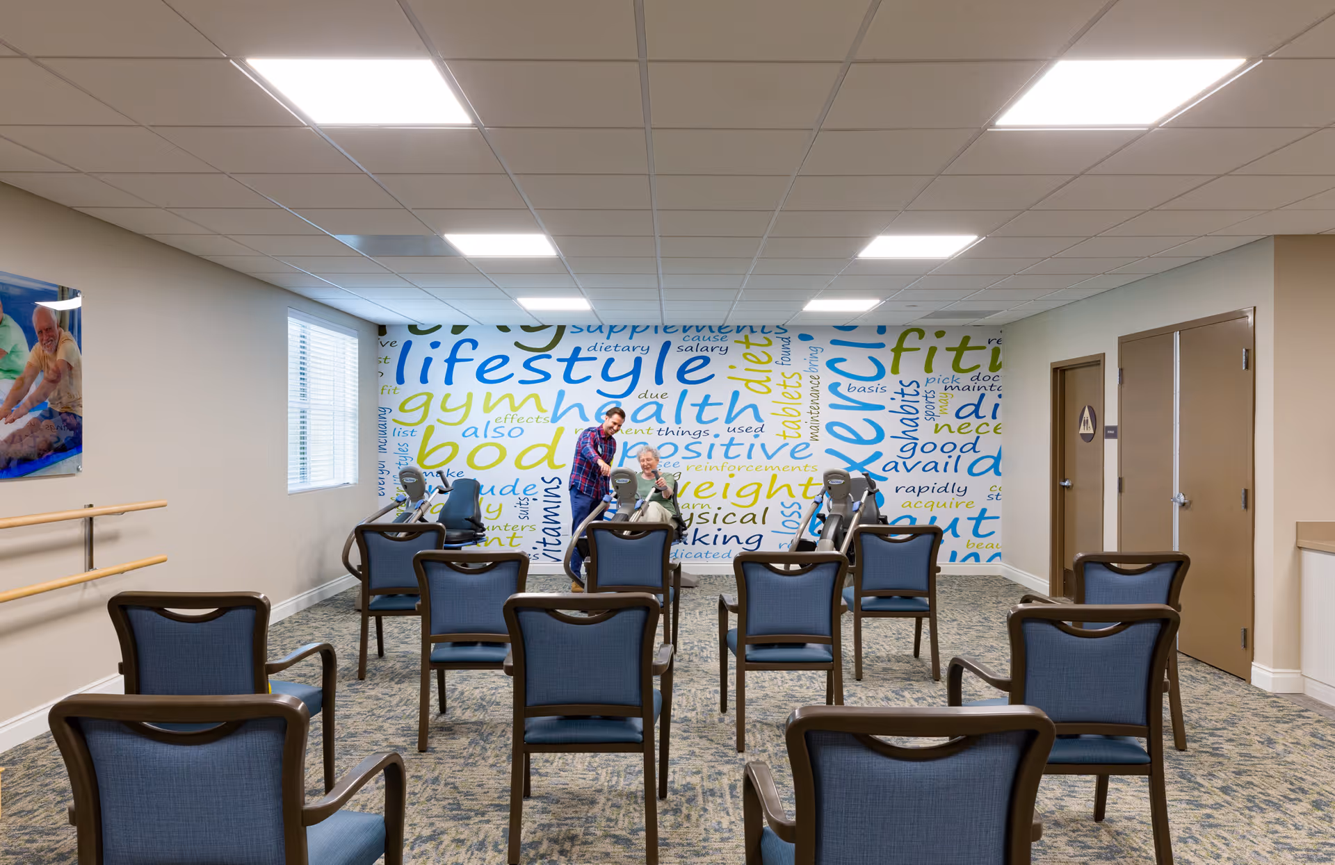 A small exercise or activity room with several chairs arranged in rows facing a wall with a colorful word cloud mural featuring health and fitness-related terms. Two people, one standing and one seated on an exercise bike, are interacting near the mural. The room has a carpeted floor, beige walls, a window with blinds, and a handrail along one wall.