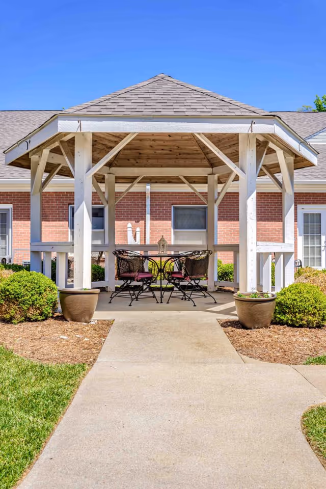Outdoor gazebo with a wooden roof and white support beams, containing a round metal table and four metal chairs with red cushions, situated on a concrete pathway surrounded by green bushes and grass, with a brick building in the background under a clear blue sky.