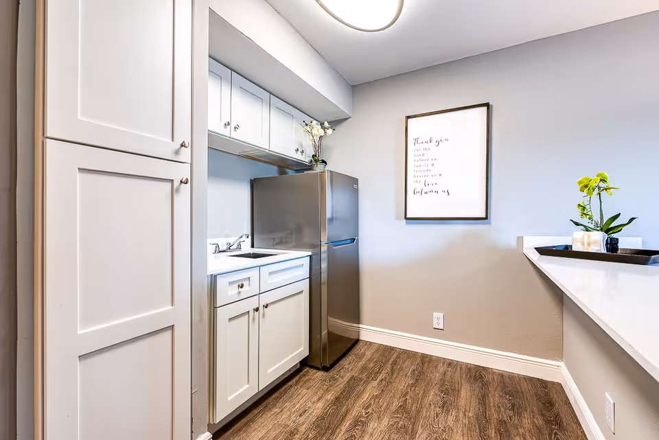 Bright kitchenette with white cabinets, a stainless steel refrigerator, sink, and a countertop with a plant and framed wall art.
