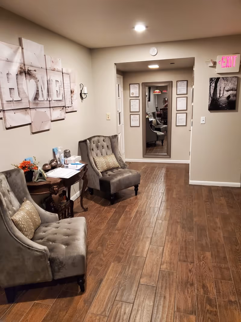 A hallway in an assisted living facility with two gray upholstered chairs with decorative pillows, a small wooden table with flowers and papers, a large wall art piece spelling 'HOME', a mirror on the far wall surrounded by framed certificates, and an illuminated exit sign above a black and white photo on the right wall. The floor is wooden and the walls are painted beige.