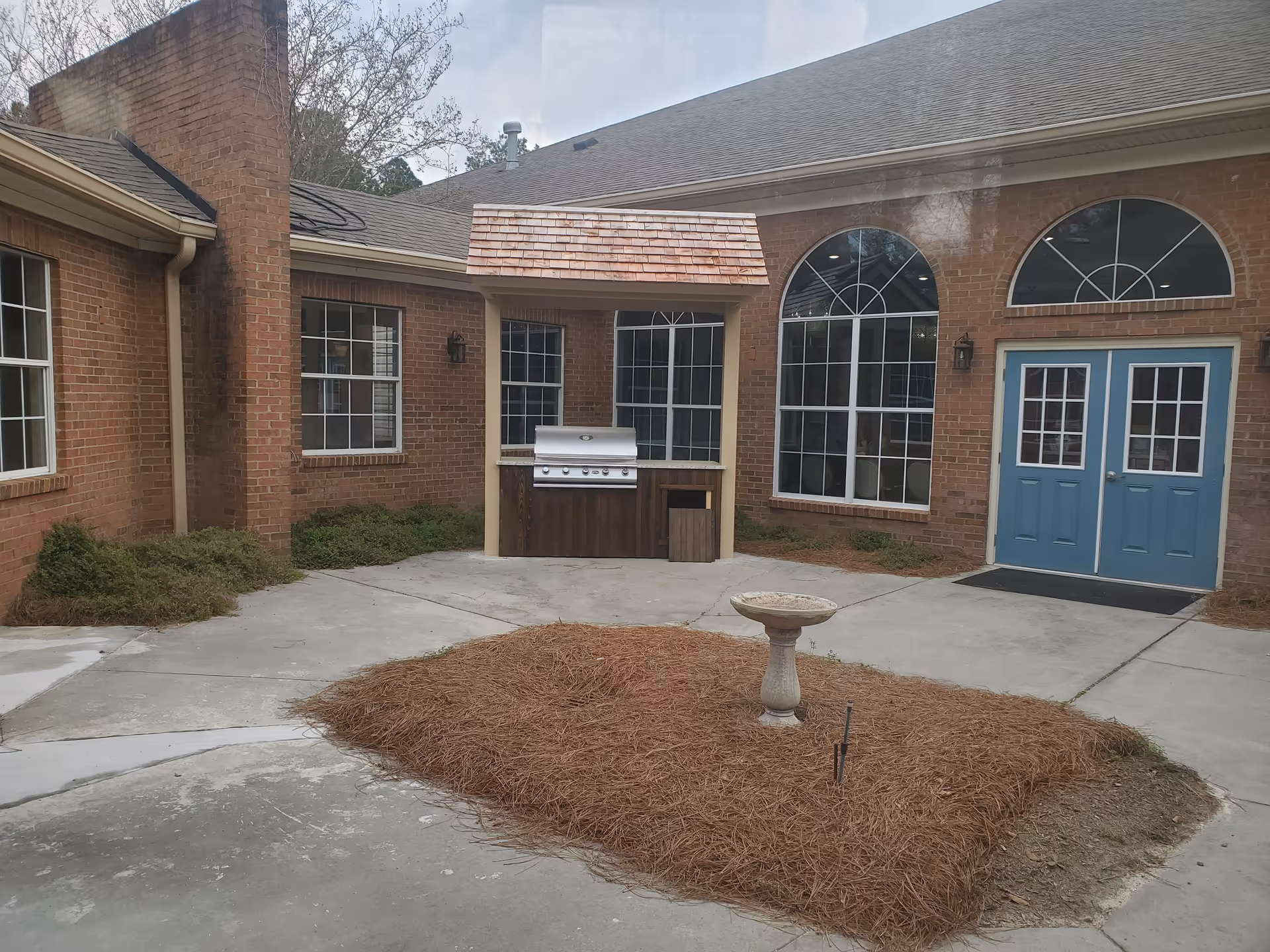 Outdoor courtyard area with a brick building featuring multiple windows and blue double doors. There is a built-in grill with a small roof above it, surrounded by concrete pavement. In the center of the courtyard is a patch of ground covered with pine straw and a birdbath.