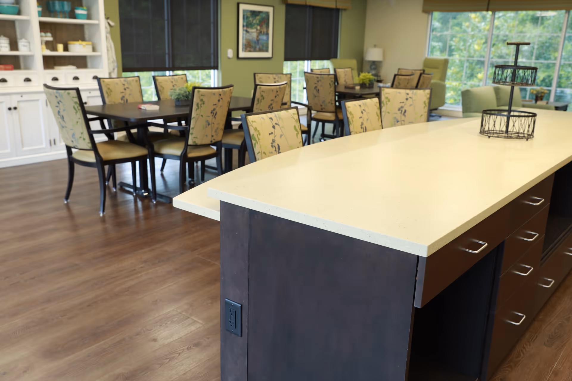 A spacious dining area with multiple tables and chairs featuring floral upholstery. The room has wooden flooring, green walls, large windows with views of greenery outside, and a white cabinet with shelves on the left. In the foreground, there is a large kitchen island with a light-colored countertop and dark cabinetry underneath.