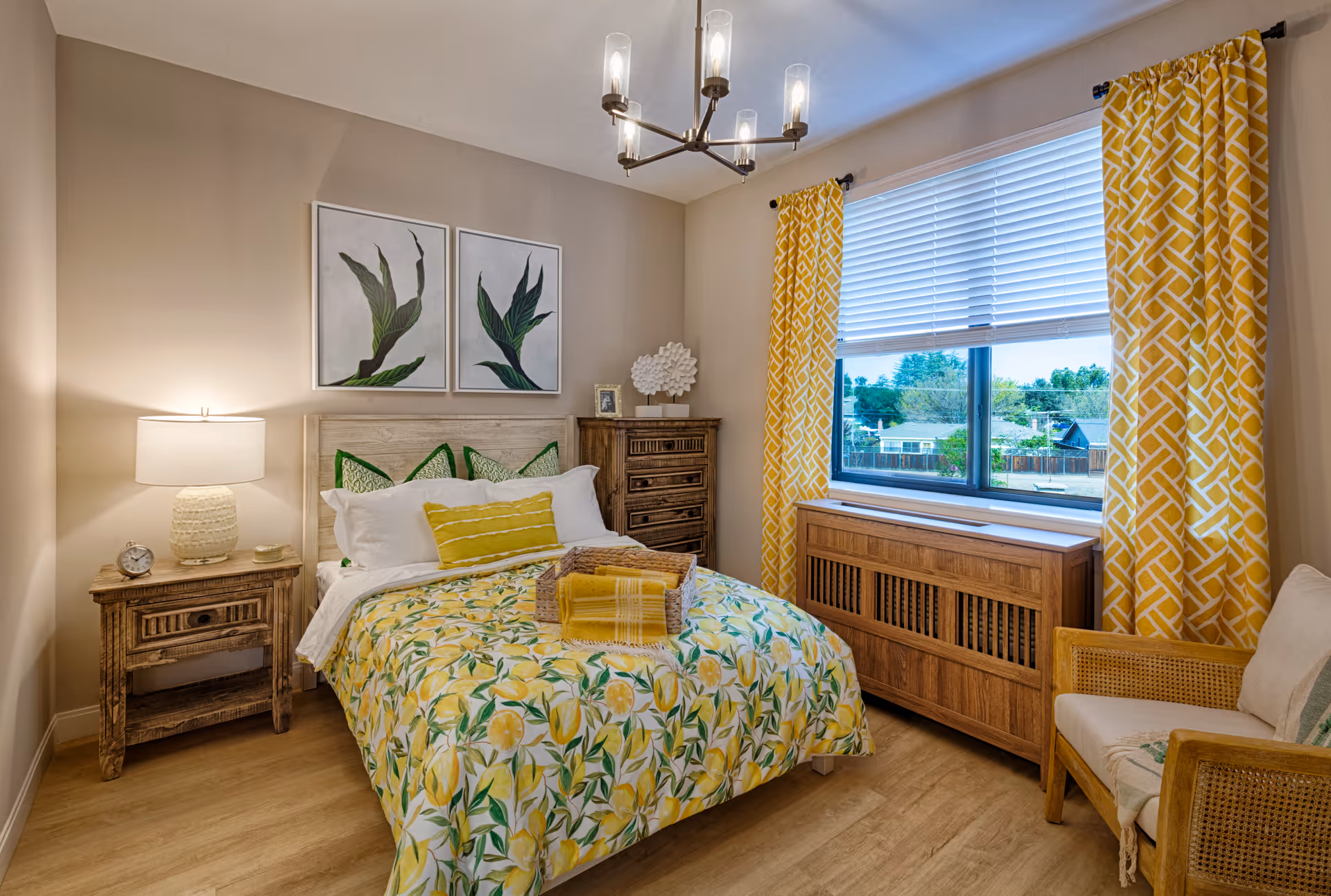 A cozy bedroom with a bed featuring a yellow and green floral bedspread, green and yellow pillows, and a folded yellow blanket on top. There is a wooden nightstand with a lamp and a clock on the left side of the bed, and a wooden dresser with decorative items on top against the back wall. Two framed botanical prints hang above the bed. A window with white blinds and yellow patterned curtains lets in natural light. A wooden chair with a cushion is positioned near the window. The room has light-colored walls and wood flooring.