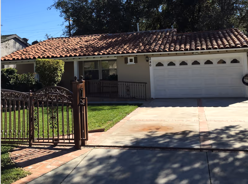 Front exterior view of a single-story residential building with a tiled roof, a white garage door, a driveway, a wrought iron gate, and a small lawn with bushes.
