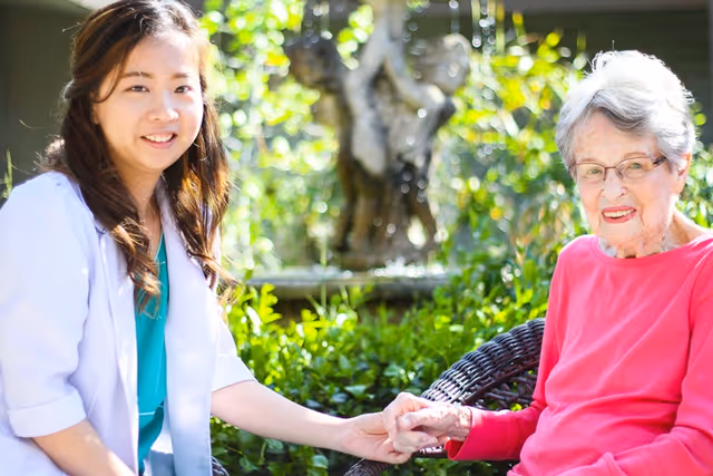 A young female healthcare worker in a white coat holding hands with an elderly woman wearing glasses and a pink sweater, sitting outdoors in a garden with greenery and a water fountain in the background.