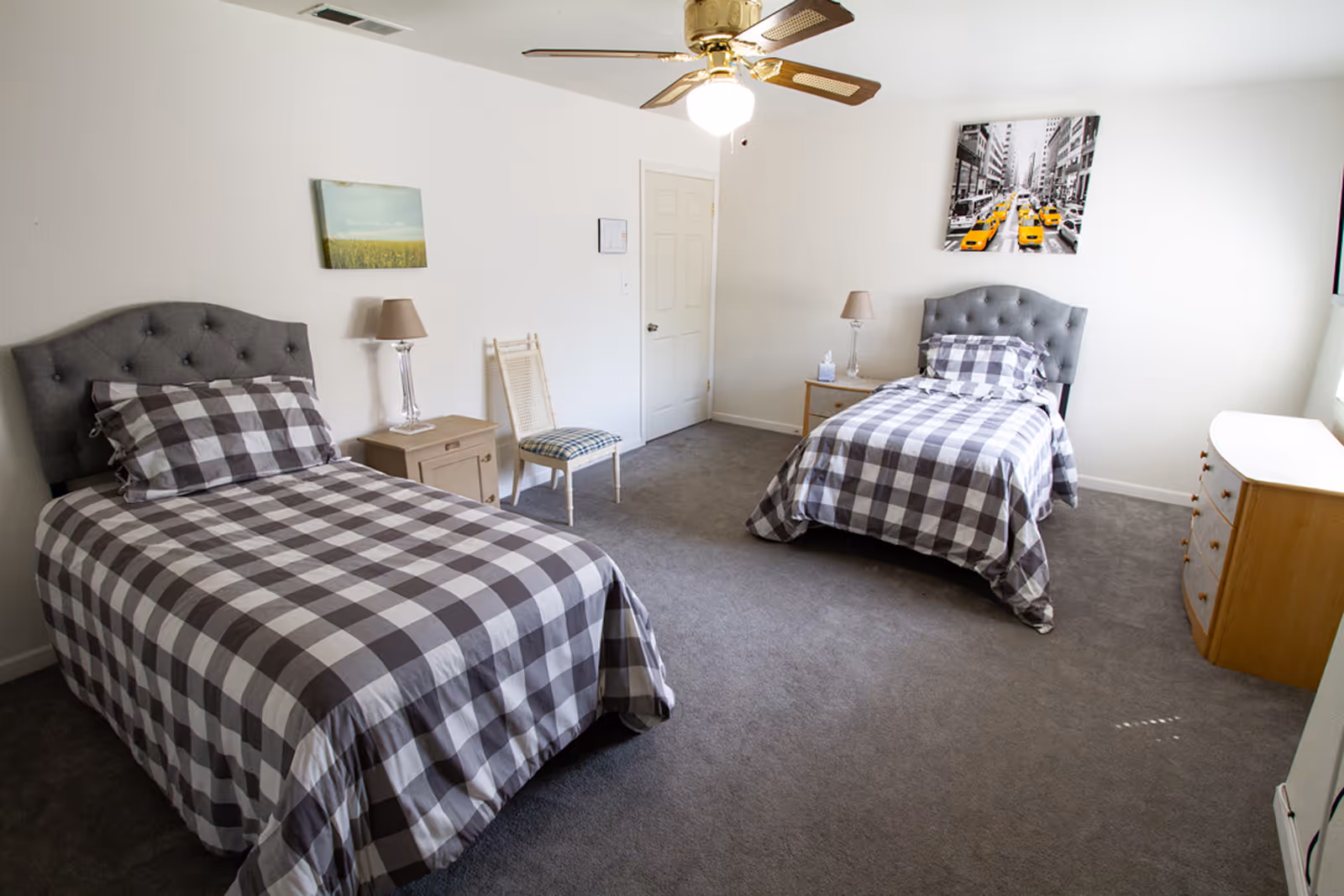Bright bedroom with two twin beds dressed in gray-and-white checkered bedding, tufted headboards, nightstands, a dresser, and a ceiling fan.