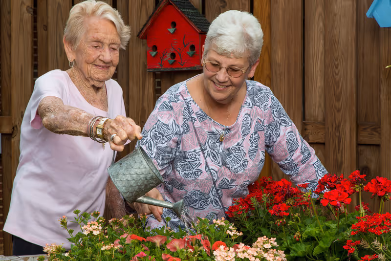 Two elderly women gardening together outdoors, one watering flowers with a metal watering can while the other smiles and watches. They are surrounded by vibrant red and pink flowers with a wooden fence and birdhouses in the background.