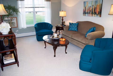 A cozy living room with a beige sofa adorned with blue patterned pillows, two teal armchairs, a wooden coffee table with decorative items, a side table with a lamp, a large window with white curtains letting in natural light, a potted plant, and a colorful abstract painting on the wall.