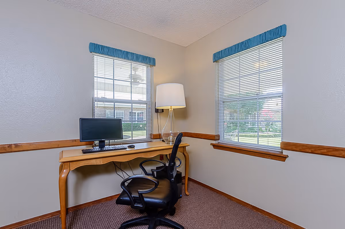 Small office nook with a wooden desk, computer monitor, black office chair, and lamp between two windows.