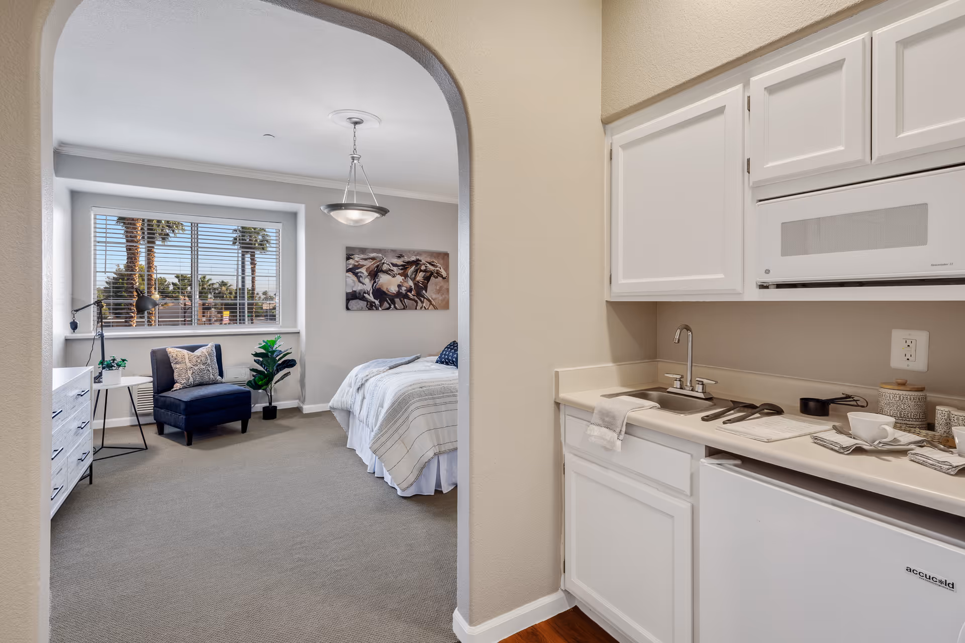 View through an arched doorway showing a small kitchenette with white cabinets, a sink, microwave, and mini fridge on the right. Beyond the kitchenette is a bedroom area with a bed, a dark blue armchair, a white dresser, a side table with a lamp, and a large window with blinds revealing palm trees outside. A painting of running horses hangs on the bedroom wall.
