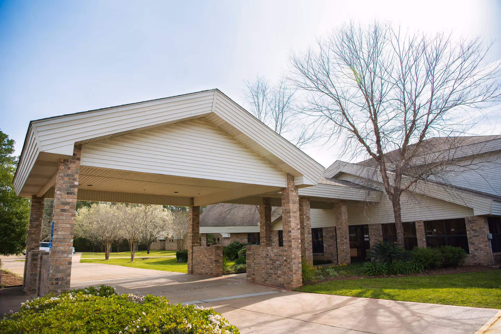 Exterior view of The Oaks Center showing a covered entrance supported by brick pillars, with a driveway and landscaped greenery including trees and bushes under a clear sky.