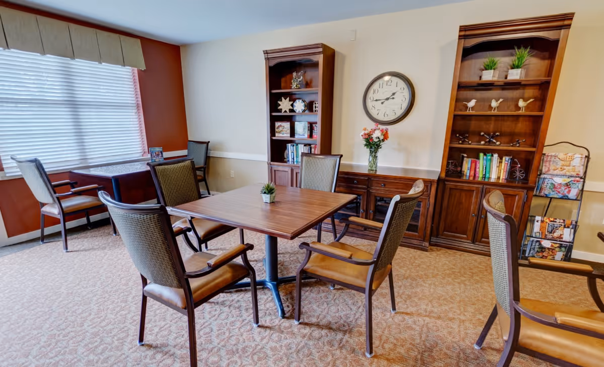 Cozy common room with a square table surrounded by chairs, bookshelves, a wall clock, and a window with blinds.