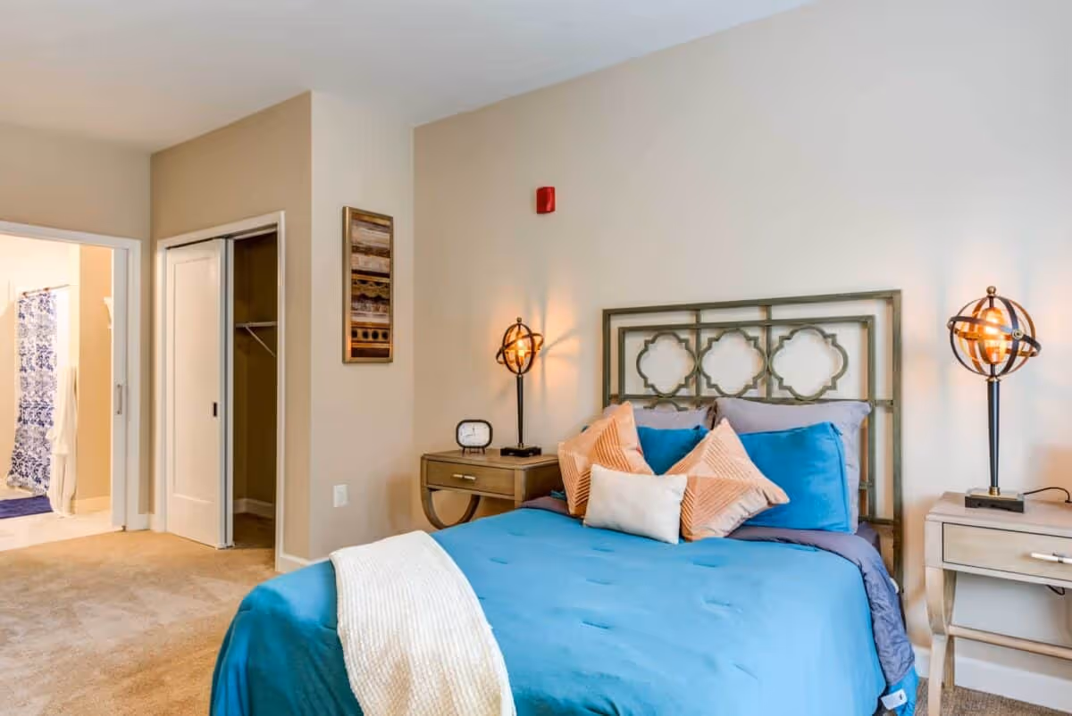 A bedroom with a blue bedspread and multiple pillows on the bed. There are two bedside tables with decorative lamps on each side of the bed. The room has beige walls and carpeted floor. An open closet with sliding doors and a bathroom with a blue and white shower curtain are visible in the background.