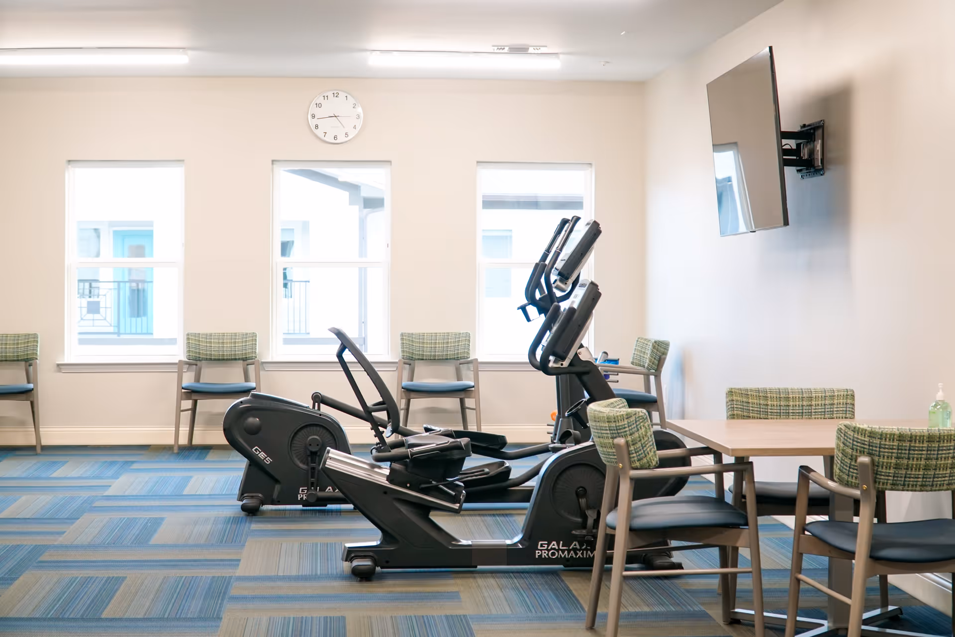 A bright room with exercise equipment including stationary bikes and elliptical machines, several green plaid chairs, a table, a wall-mounted TV, and three windows letting in natural light. A clock is mounted on the wall above the windows.
