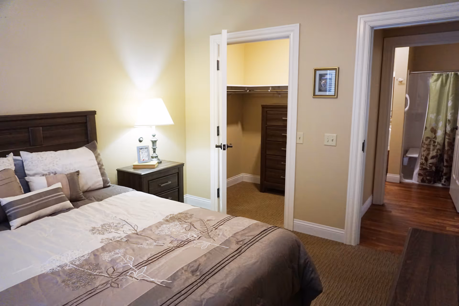 A cozy bedroom in Snyder Village Assisted Living featuring a neatly made bed with multiple pillows and a floral-patterned comforter. A wooden nightstand with a lamp, a framed photo, and a book sits beside the bed. There is an open door leading to a walk-in closet with a wooden dresser inside, and another open door reveals a bathroom with a shower curtain and wooden flooring.