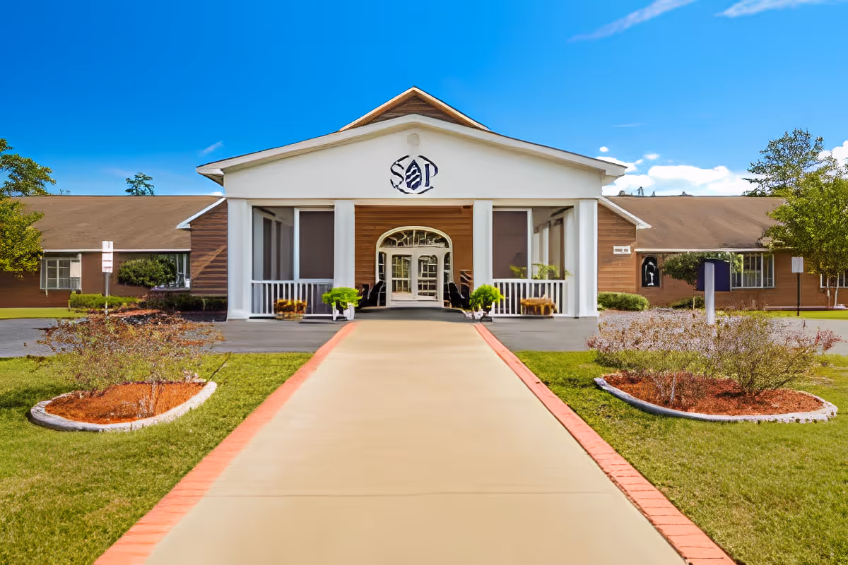 Front exterior view of Southern Pines Senior Living facility with a wide concrete walkway leading to the entrance, flanked by green lawns and landscaped flower beds. The building has a white portico with columns and a brown roof under a clear blue sky.