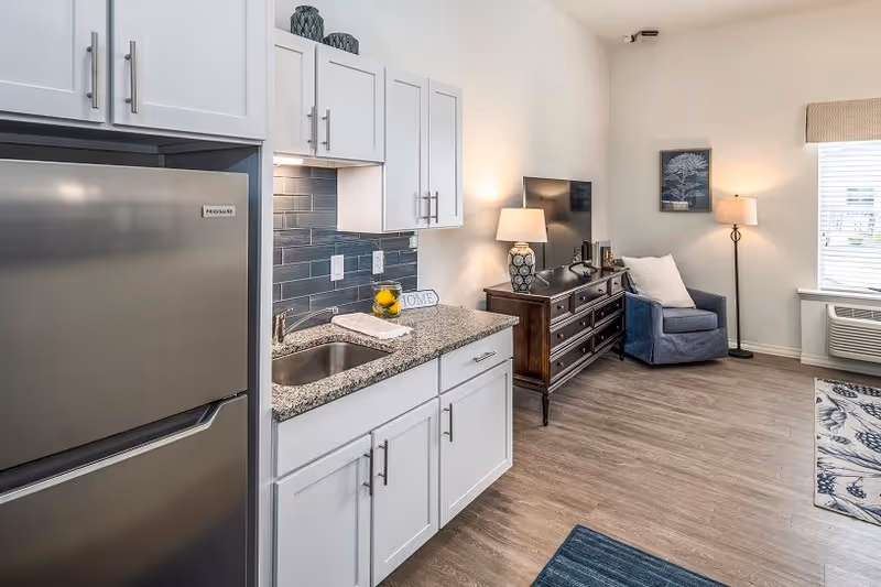 A modern living space featuring a small kitchen area with white cabinets, a stainless steel refrigerator, and a granite countertop with a sink. Adjacent to the kitchen is a living area with a dark wooden dresser, a flat-screen TV, a blue armchair with a white pillow, a floor lamp, and a window with blinds. The room has light-colored walls and wood flooring.