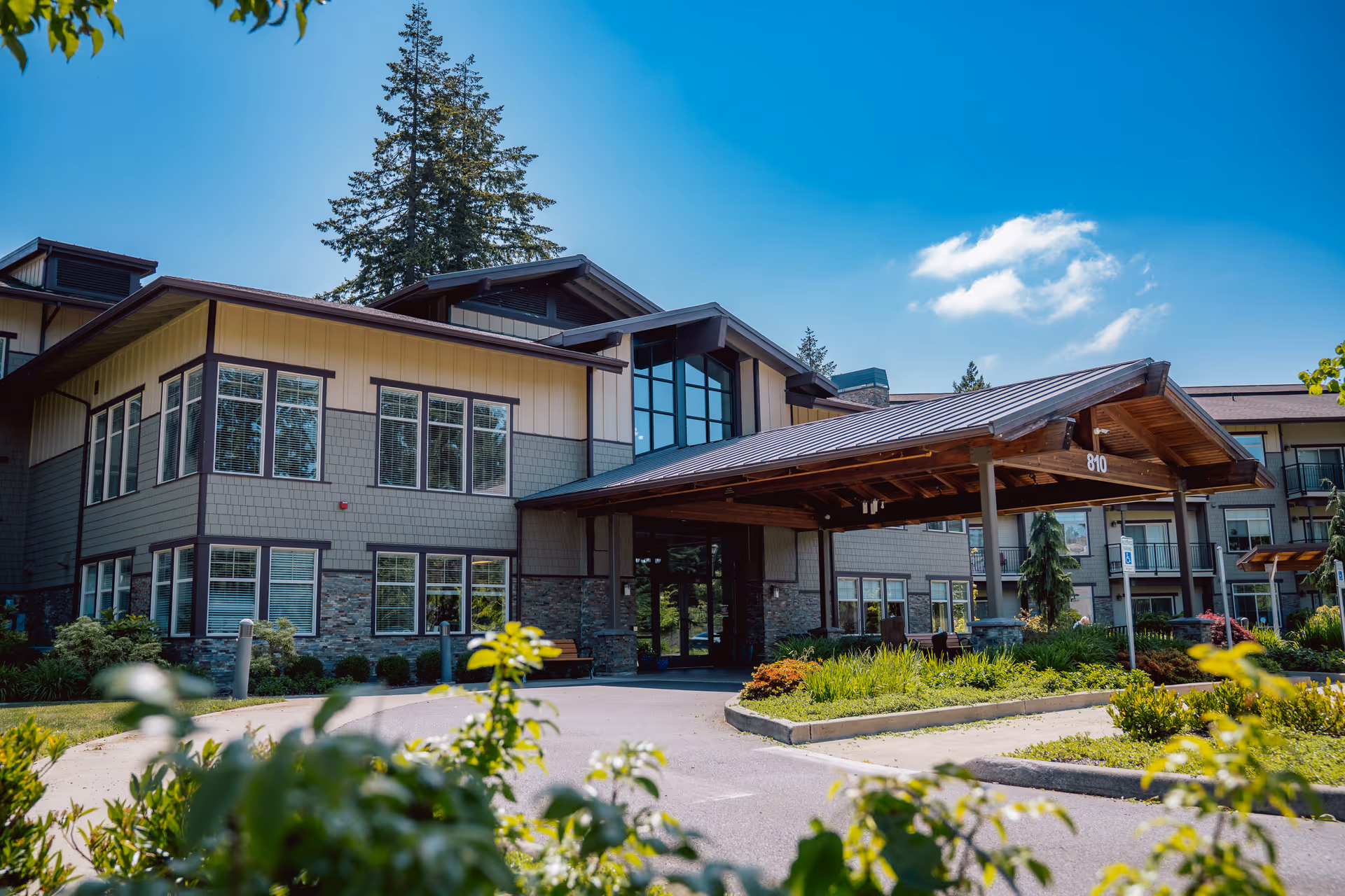 Exterior front entrance of a multi-story senior living building with a covered porte-cochere, landscaped driveway, and large windows.