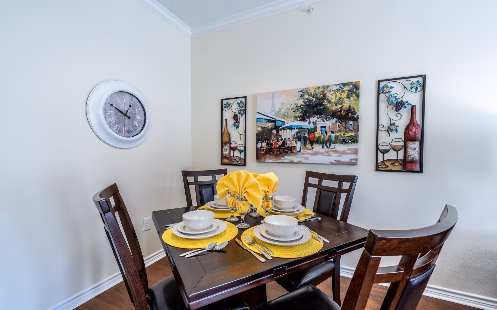 A dining area with a dark wooden table set for four people. Each place setting includes a white bowl, plate, silverware, and a yellow placemat. Yellow cloth napkins are folded in the center of the table. On the wall behind the table, there is a large painting depicting an outdoor café scene and two decorative wall hangings featuring wine bottles and glasses. A round wall clock with Roman numerals is mounted on the adjacent wall.