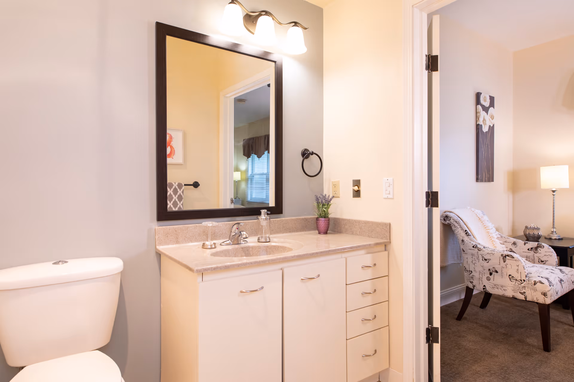 Bright bathroom with a vanity, sink and mirror, a toilet to the left, and an open doorway revealing a nearby sitting area.