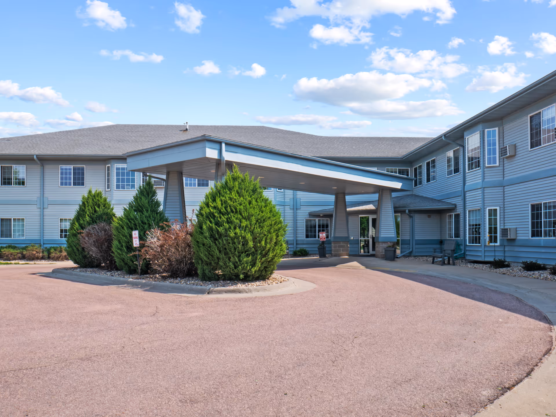 Exterior view of Good Samaritan Society - Pipestone - Ridge View Estates building with a covered entrance, surrounded by shrubs and a clear blue sky with scattered clouds.