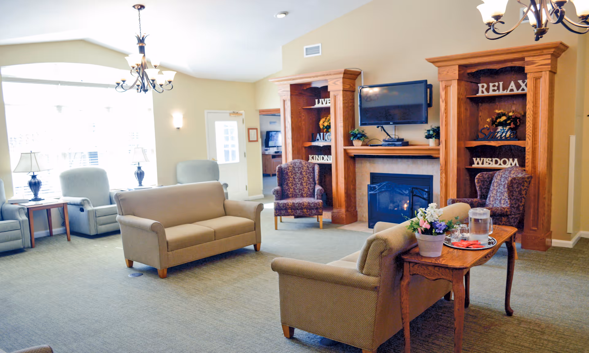 Sunny assisted-living lounge with sofas and armchairs arranged around a fireplace and wall-mounted TV.