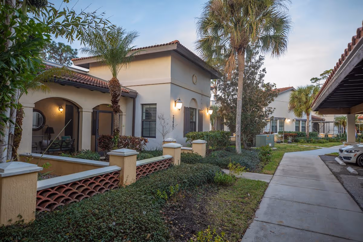 Landscaped courtyard and front walkway of a Mediterranean-style senior living building with palm trees, entry porches, and covered parking.