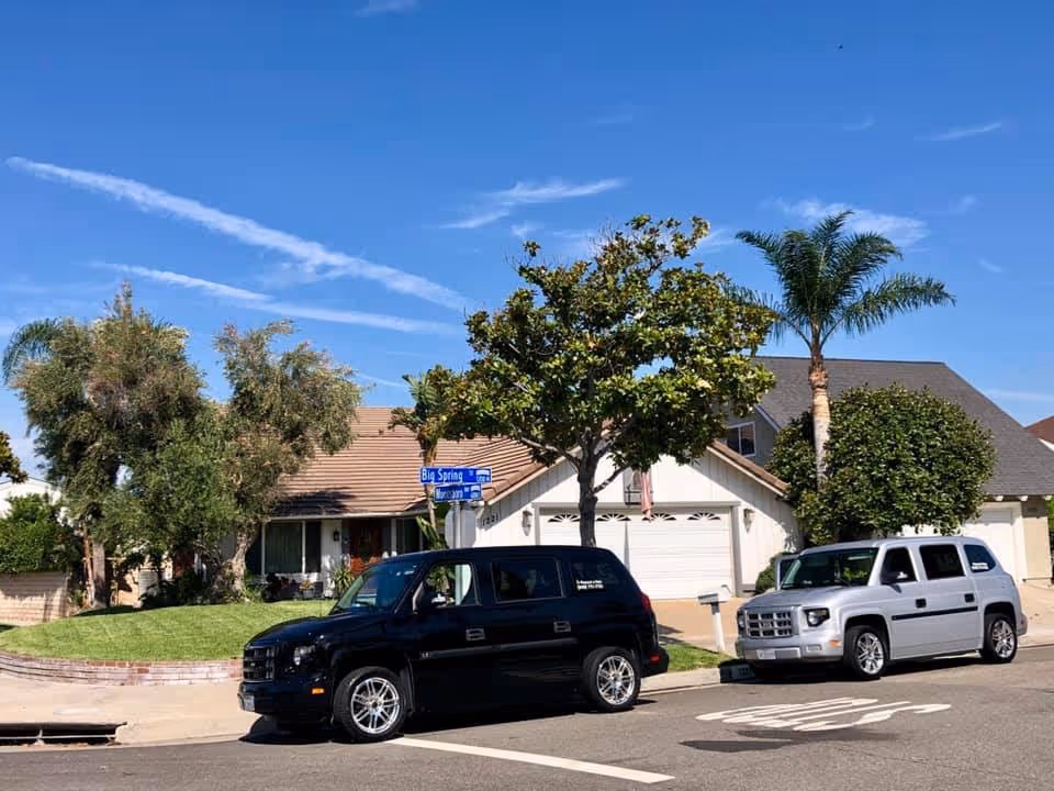 Street view of a residential neighborhood with two parked cars, a black SUV and a silver SUV, in front of a house with a garage. The house is surrounded by trees and greenery under a clear blue sky.