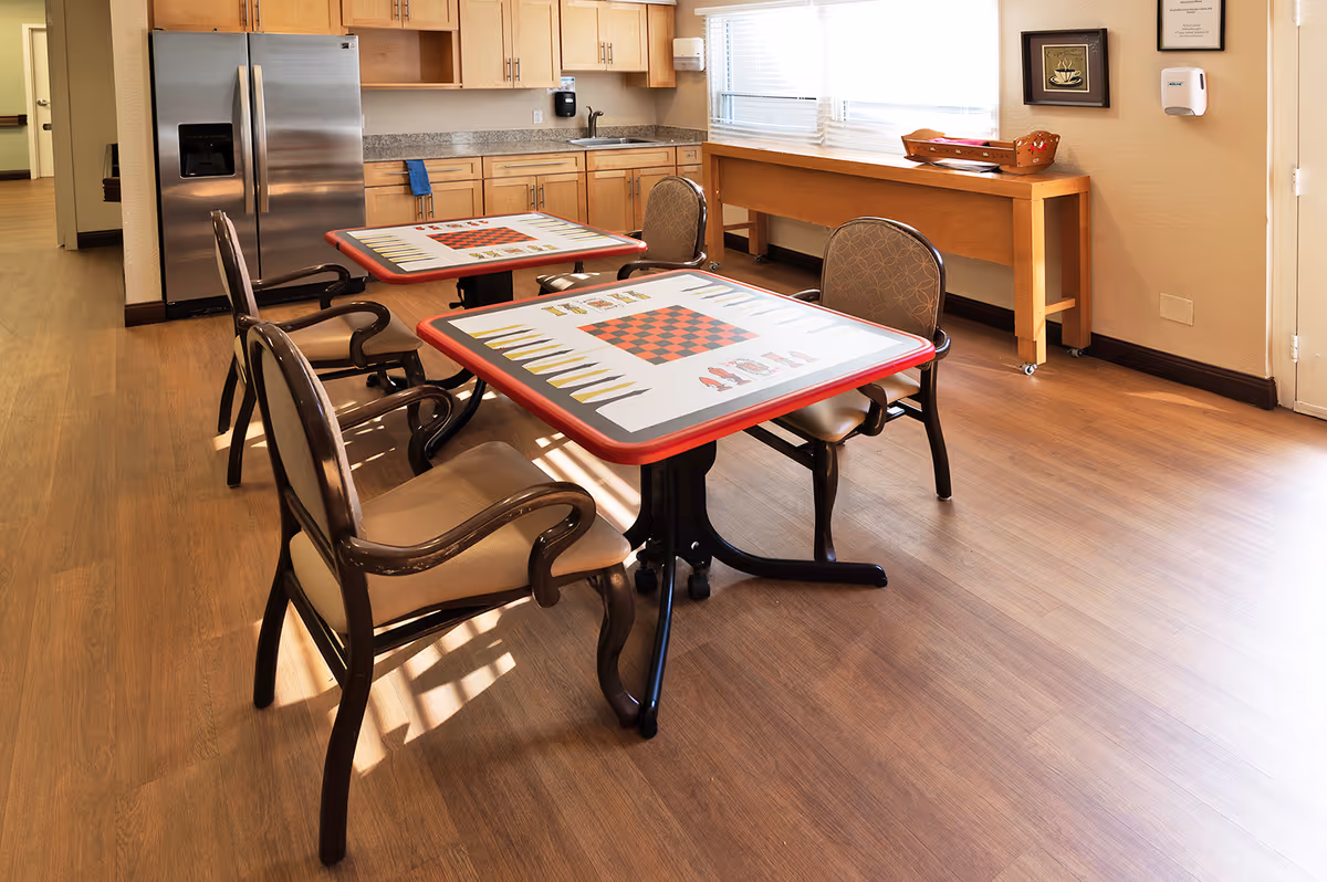 A bright room with two tables featuring backgammon and chess game boards on their surfaces, surrounded by four cushioned wooden chairs. The room has wooden flooring, a stainless steel refrigerator, wooden cabinets, a sink, and a window with blinds letting in natural light. A wooden console table is placed against the wall with a basket on top.