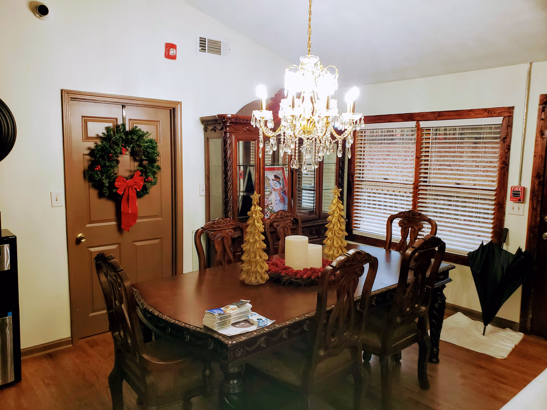 Ornate wooden dining room with a chandelier above a decorated table holding candles and gold tree decorations, a wreath on a closed door, and a china cabinet.