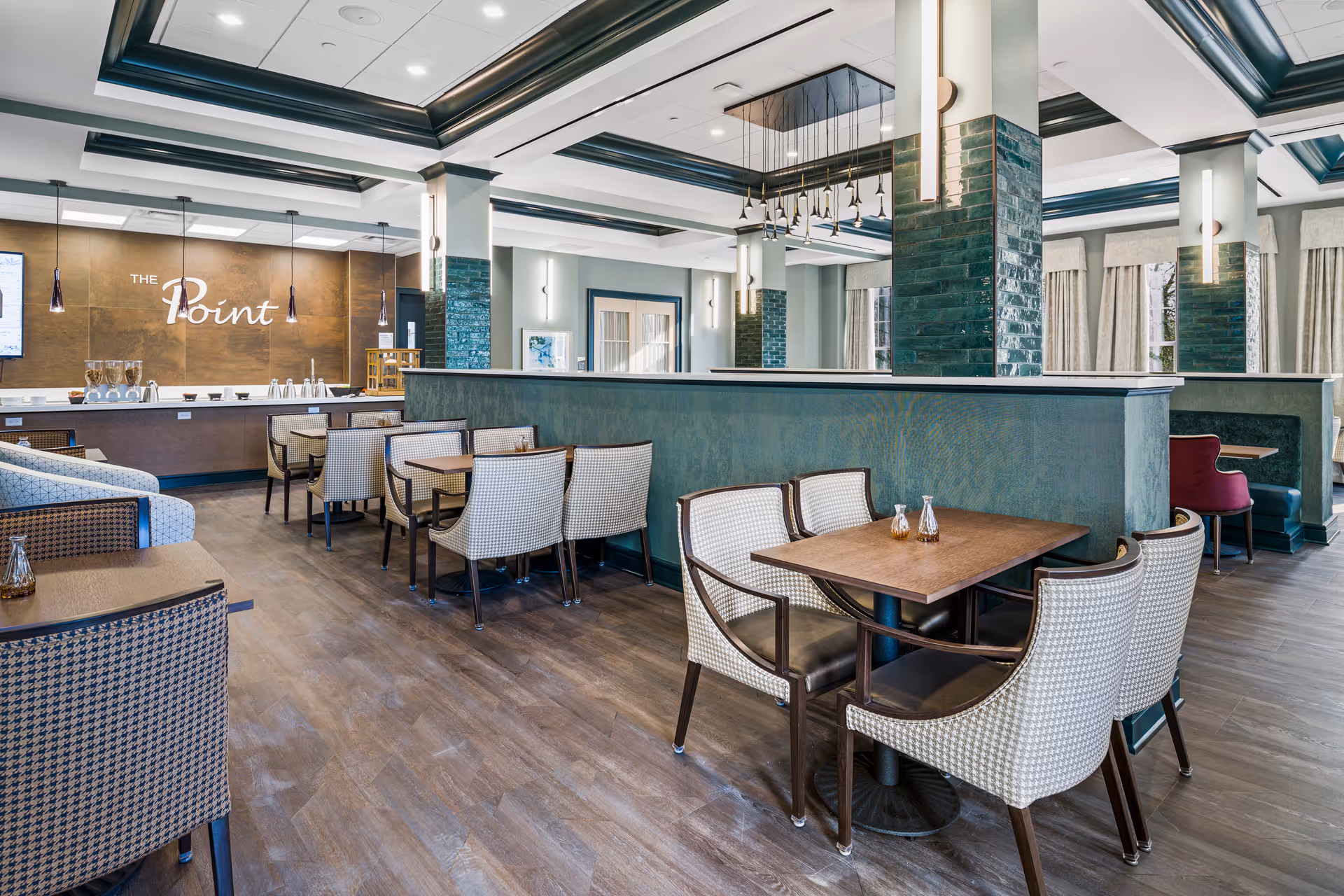 A spacious, well-lit dining area with multiple tables and cushioned chairs arranged neatly. The room features green tiled pillars, large windows with curtains, and a serving counter in the background with the words 'The Point' displayed on the wall. The flooring is wood-style, and modern light fixtures hang from the ceiling.