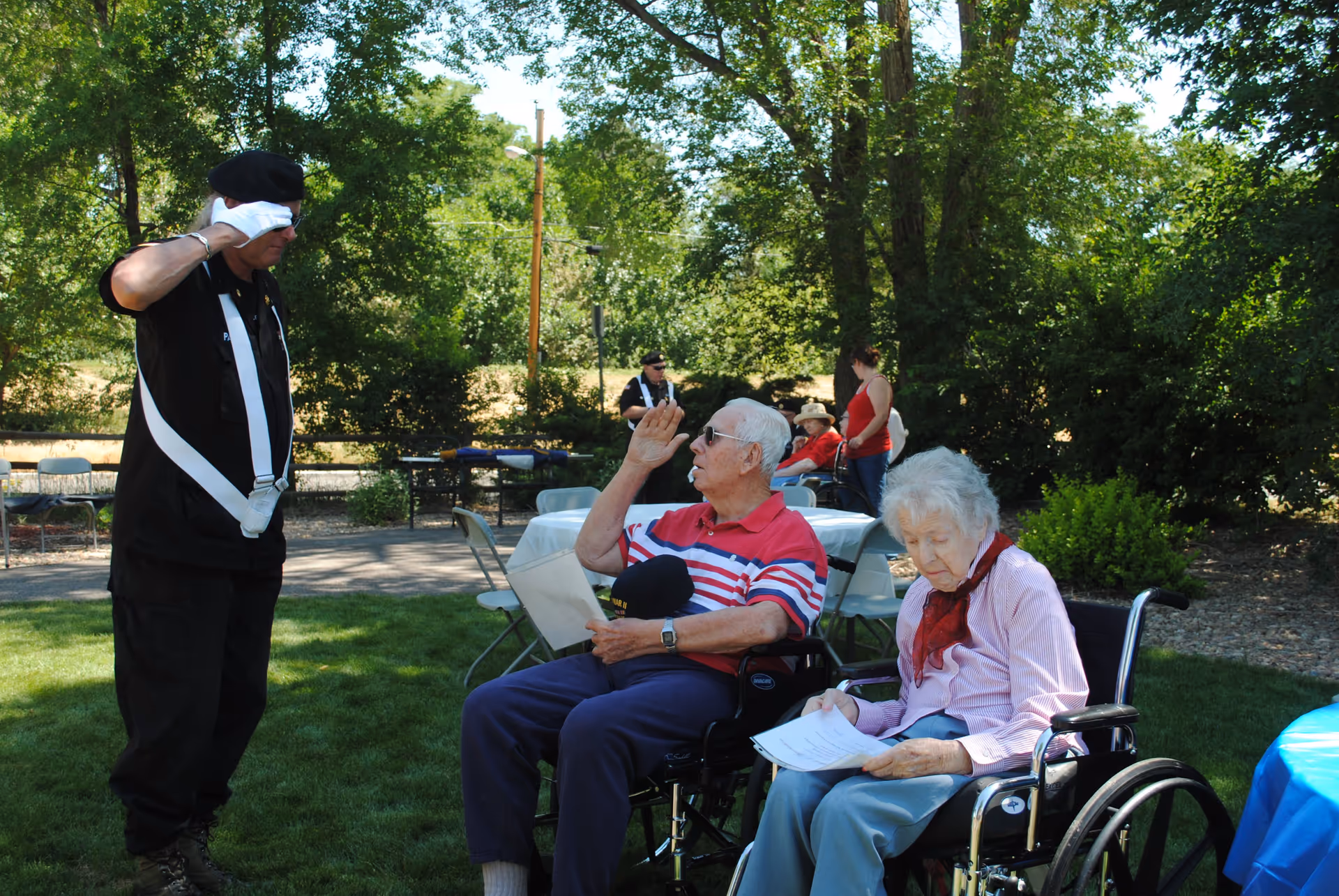 An elderly man and woman in wheelchairs outdoors on a grassy area. The man is saluting a person dressed in black with a beret and white gloves. Other people and tables with chairs are visible in the background surrounded by trees and greenery.